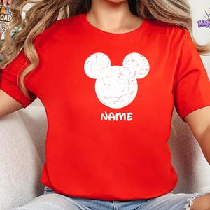 May include: A bright red short-sleeved t-shirt featuring a large, white, distressed-texture Mickey Mouse head silhouette centered on the chest. Below the graphic, the word NAME is printed in a bold, white, sans-serif font.