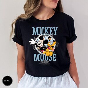 May include: Black t-shirt featuring a graphic of Mickey Mouse and Pluto. The design includes the words "Mickey Mouse" in blue, with Mickey Mouse waving and Pluto sitting. The shirt is a classic crew neck.