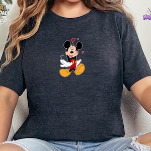 May include: A dark heather gray short-sleeve t-shirt featuring a centered graphic of Mickey Mouse with white gloves, red shorts, and yellow shoes, surrounded by small red hearts. The character has a joyful expression with eyes closed and a smile.