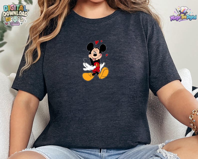 May include: A dark heather gray short-sleeve t-shirt featuring a centered graphic of Mickey Mouse with white gloves, red shorts, and yellow shoes, surrounded by small red hearts. The character has a joyful expression with eyes closed and a smile.