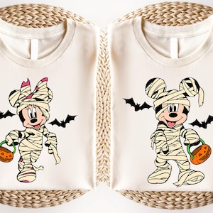 May include: Two cream-colored t-shirts featuring Halloween-themed cartoon characters. One shirt displays Minnie Mouse as a mummy with a pink bow, holding a jack-o'-lantern. The other shirt shows Mickey Mouse as a mummy, also with a jack-o'-lantern. Black bats are included.