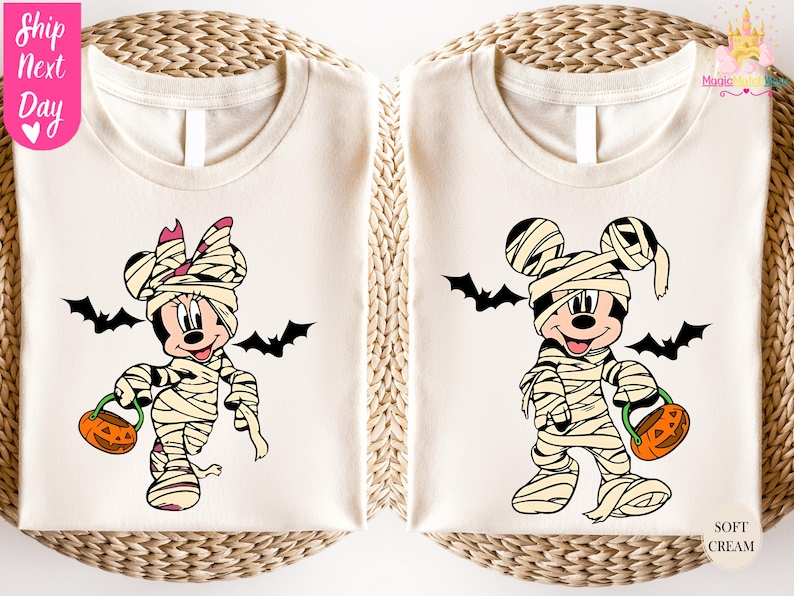 May include: Two cream-colored t-shirts featuring Halloween-themed cartoon characters. One shirt displays Minnie Mouse as a mummy with a pink bow, holding a jack-o'-lantern. The other shirt shows Mickey Mouse as a mummy, also with a jack-o'-lantern. Black bats are included.