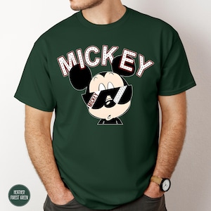 May include: A dark green t-shirt featuring a graphic of Mickey Mouse wearing sunglasses. The word "MICKEY" is printed above the character in white letters with red accents. The shirt has short sleeves and a crew neck.