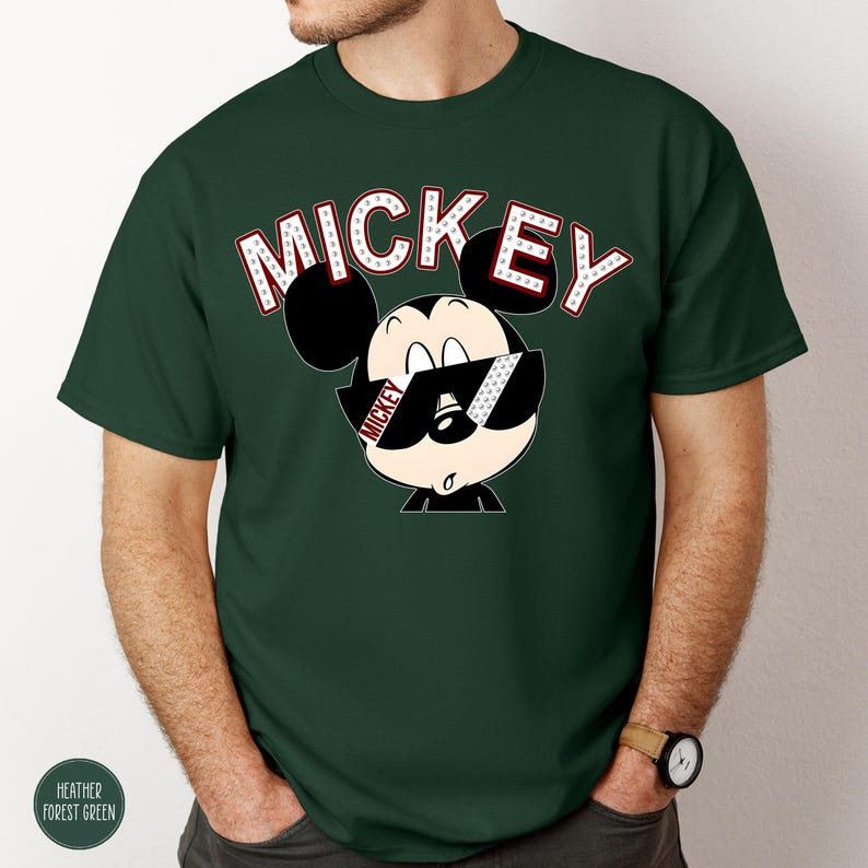 May include: A dark green t-shirt featuring a graphic of Mickey Mouse wearing sunglasses. The word "MICKEY" is printed above the character in white letters with red accents. The shirt has short sleeves and a crew neck.