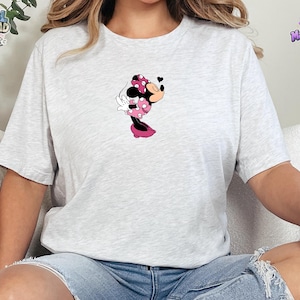 May include: A light gray heathered short-sleeve t-shirt featuring a centered graphic of a cartoon mouse character wearing a pink polka-dot dress and bow, blowing a kiss with a small black heart icon floating near the character's face.