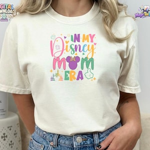 May include: A cream-colored short-sleeve t-shirt featuring a colorful graphic print on the chest. The text reads IN MY Disney MOM ERA, accompanied by a purple mouse ear silhouette with a polka dot bow, a castle icon, and a hand gesture icon.