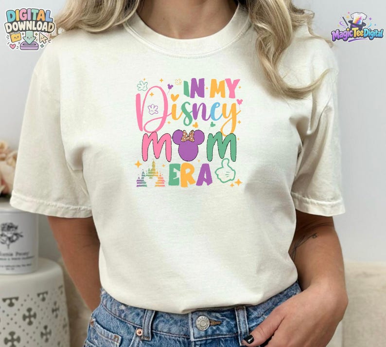 May include: A cream-colored short-sleeve t-shirt featuring a colorful graphic print on the chest. The text reads IN MY Disney MOM ERA, accompanied by a purple mouse ear silhouette with a polka dot bow, a castle icon, and a hand gesture icon.
