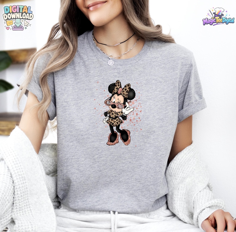 May include: A heather grey short-sleeve t-shirt featuring a graphic of a cartoon mouse character wearing leopard print clothing and pink glittery sunglasses, with a subtle pink paint splatter effect surrounding the character design.