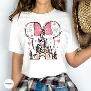 Puede incluir: Camiseta blanca con una ilustraci&oacute;n caprichosa de un castillo, un lazo rosa con lunares y orejas de Mickey Mouse. El dise&ntilde;o incluye corazones rosas y estrellas. La palabra "WHITE" est&aacute; en la parte inferior izquierda.