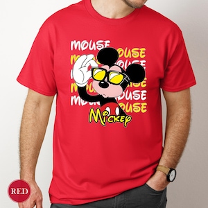 May include: Red t-shirt featuring a cartoon Mickey Mouse character wearing yellow sunglasses. The design includes the word "MOUSE" repeated in a stacked pattern, with "Mickey" in yellow script. The shirt is short-sleeved.