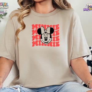May include: A beige short-sleeved t-shirt featuring a graphic print of the character Minnie Mouse in black and white. Behind the character, the word MINNIE is repeated in a wavy, red, stacked font pattern across the center of the chest.