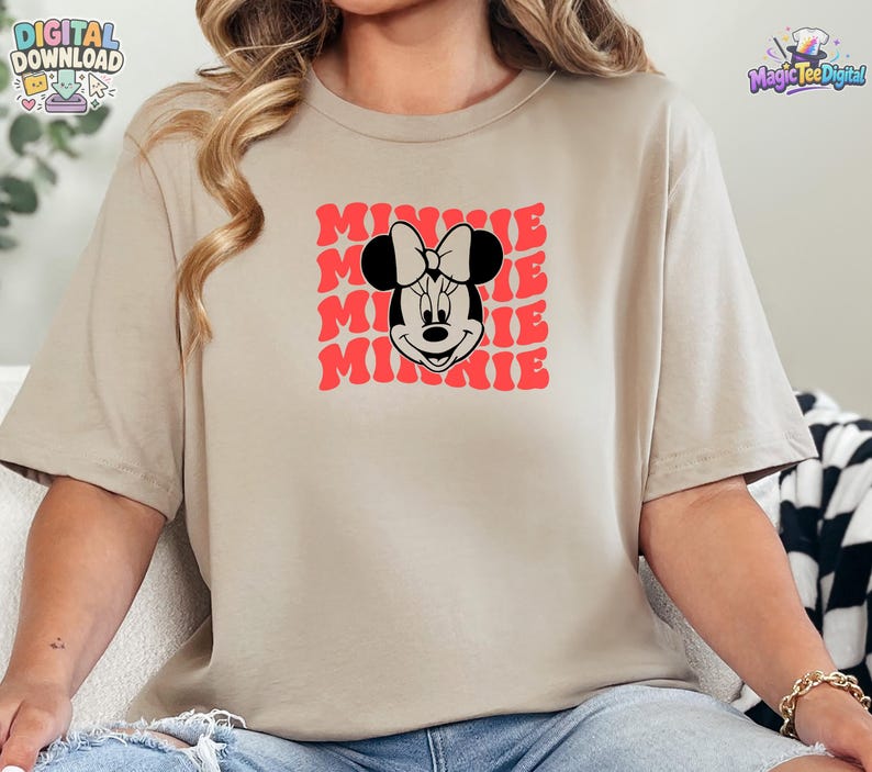May include: A beige short-sleeved t-shirt featuring a graphic print of the character Minnie Mouse in black and white. Behind the character, the word MINNIE is repeated in a wavy, red, stacked font pattern across the center of the chest.