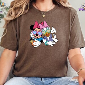 May include: A brown short-sleeved t-shirt featuring a graphic print of Minnie Mouse and Daisy Duck wearing heart-shaped sunglasses. Minnie holds an orange drink with a slice of lemon, while Daisy holds a white ice cream cone. Both characters wear pink bows.