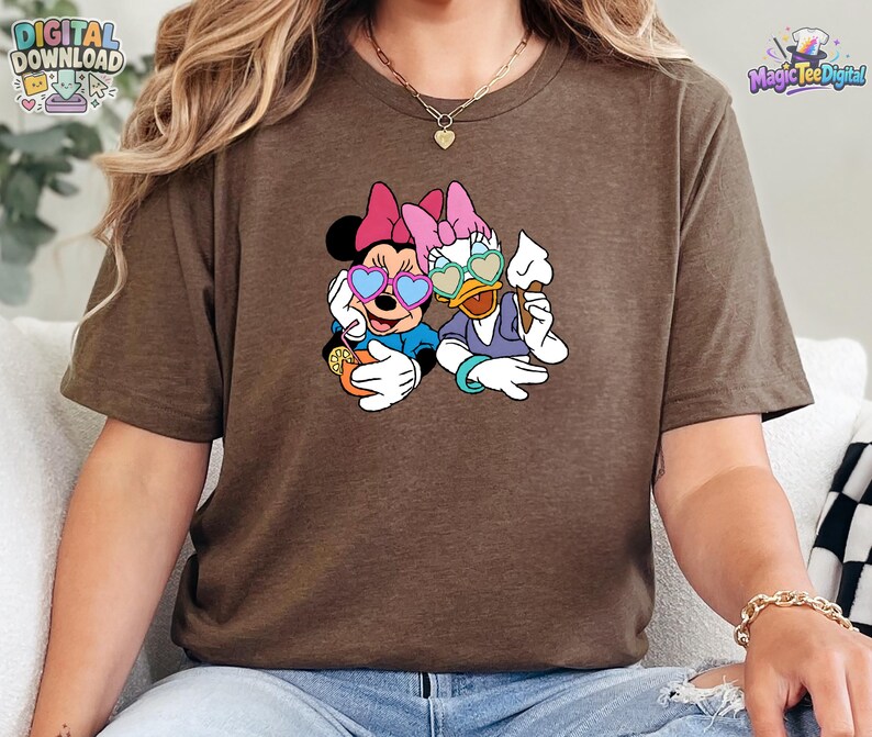 May include: A brown short-sleeved t-shirt featuring a graphic print of Minnie Mouse and Daisy Duck wearing heart-shaped sunglasses. Minnie holds an orange drink with a slice of lemon, while Daisy holds a white ice cream cone. Both characters wear pink bows.