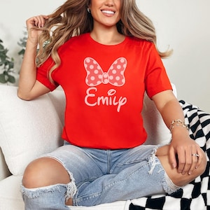 May include: A red t-shirt featuring a white polka-dot bow and the name "Emily" in a script font. The shirt is a crew neck style and is made of a soft material. The shirt is a digital download.