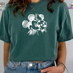 May include: A dark green short-sleeved t-shirt featuring a white graphic print of two cartoon mouse characters on the chest. The design is rendered in a sketch-like style with visible line work. The shirt is paired with blue denim jeans.