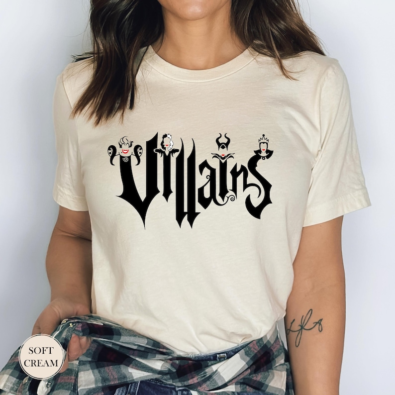 May include: Cream-colored t-shirt featuring the word "Villains" in a gothic font, with stylized character illustrations above the letters. The shirt has short sleeves and a crew neck.