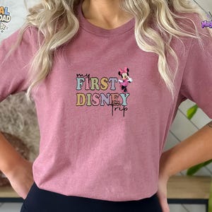May include: A mauve short-sleeved t-shirt featuring a graphic print with the text "MY FIRST DISNEY TRIP" in colourful, retro-style bubble lettering. A small illustration of Minnie Mouse wearing a pink dress and bow is positioned above the text.