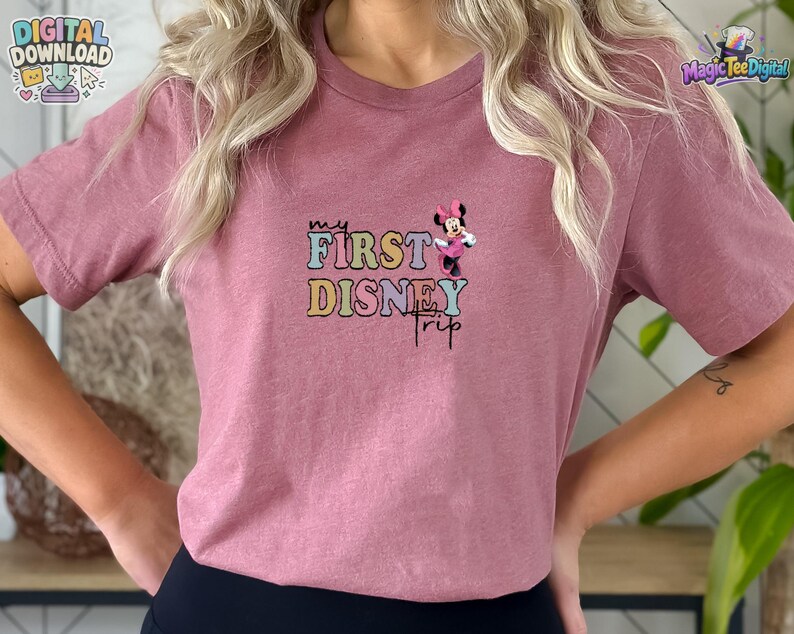 May include: A mauve short-sleeved t-shirt featuring a graphic print with the text "MY FIRST DISNEY TRIP" in colourful, retro-style bubble lettering. A small illustration of Minnie Mouse wearing a pink dress and bow is positioned above the text.