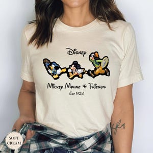 May include: Cream-colored t-shirt featuring a graphic of Disney characters Donald Duck, Mickey Mouse, and Goofy. The text "Disney Mickey Mouse & Friends Est 1928" is printed in black script. The shirt has short sleeves.