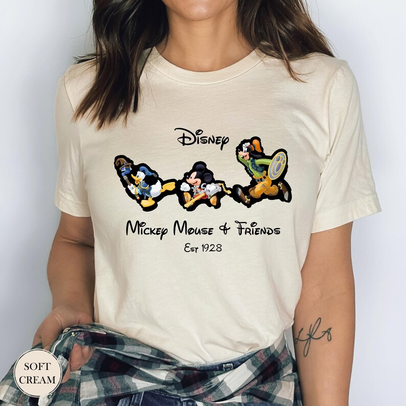 May include: Cream-colored t-shirt featuring a graphic of Disney characters Donald Duck, Mickey Mouse, and Goofy. The text "Disney Mickey Mouse & Friends Est 1928" is printed in black script. The shirt has short sleeves.