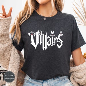 May include: Dark heather gray t-shirt with the word "Villains" in a stylized white font. The word is decorated with small, themed graphics above each letter. The shirt is a crew neck style.