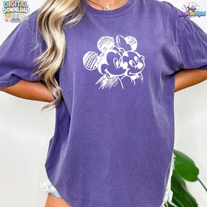 May include: A purple short-sleeved t-shirt featuring a white line-art graphic of Mickey Mouse and Minnie Mouse on the chest. The shirt has a crew neckline and a relaxed fit, with the printed design showing the two characters side by side.