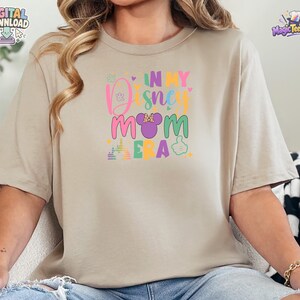 May include: A beige short-sleeved t-shirt featuring a colorful graphic print on the chest. The text reads IN MY DISNEY MOM ERA, accompanied by a purple mouse ear silhouette with a polka dot bow, a castle outline, and small heart icons.
