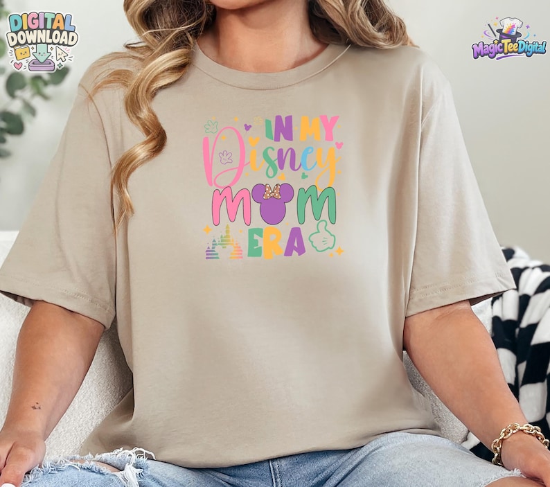 May include: A beige short-sleeved t-shirt featuring a colorful graphic print on the chest. The text reads IN MY DISNEY MOM ERA, accompanied by a purple mouse ear silhouette with a polka dot bow, a castle outline, and small heart icons.