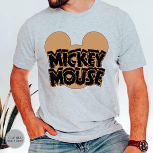 Puede incluir: Camiseta gris claro con un dise&ntilde;o de cabeza de Mickey Mouse marr&oacute;n y las palabras "MICKEY MOUSE" en un estampado de leopardo negro y marr&oacute;n. La camiseta est&aacute; hecha de un material suave.