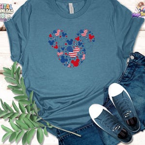 Puede incluir: Una camiseta de cuello redondo en color azul jaspeado con un gr&aacute;fico grande de la silueta de la cabeza de Mickey Mouse compuesta por iconos rojos, blancos y azules, incluyendo patrones de estrellas y rayas, junto a vaqueros azul oscuro y zapatillas.