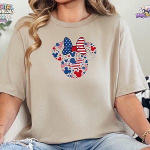 Puede incluir: Una camiseta beige de manga corta con un gr&aacute;fico central de una silueta de cabeza de rat&oacute;n compuesta por iconos m&aacute;s peque&ntilde;os en rojo, blanco y azul, incluyendo patrones de estrellas y rayas, rematada con un gran lazo de dise&ntilde;o patri&oacute;tico.