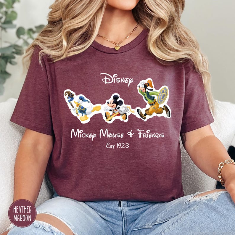 May include: Heather maroon t-shirt featuring the Disney logo and the phrase "Mickey Mouse & Friends Est 1928." The shirt displays cartoon images of Mickey Mouse, Donald Duck, and Goofy. The shirt is a crew neck style.