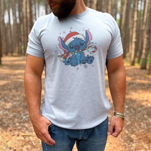 May include: Heather light grey t-shirt featuring a cartoon character wearing a Santa hat and holding a candy cane. The character is blue with large ears and is surrounded by snowflakes. The shirt is a digital download.