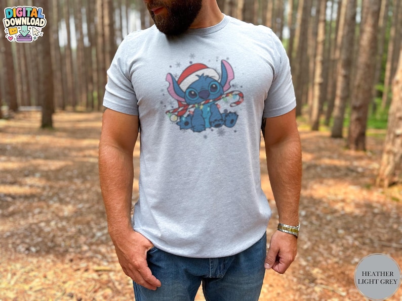 May include: Heather light grey t-shirt featuring a cartoon character wearing a Santa hat and holding a candy cane. The character is blue with large ears and is surrounded by snowflakes. The shirt is a digital download.