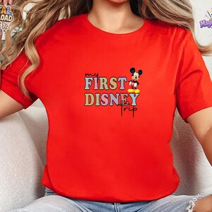 May include: A bright red short-sleeved t-shirt featuring a graphic print with the text "my FIRST DISNEY trip" in colourful, blocky letters. A small illustration of Mickey Mouse is positioned next to the word "FIRST" on the shirt.