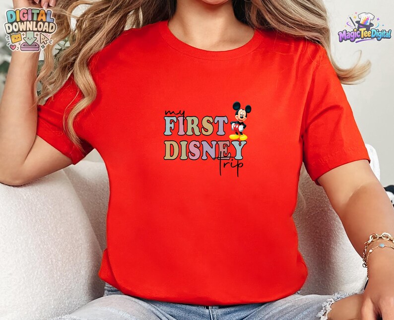 May include: A bright red short-sleeved t-shirt featuring a graphic print with the text "my FIRST DISNEY trip" in colourful, blocky letters. A small illustration of Mickey Mouse is positioned next to the word "FIRST" on the shirt.