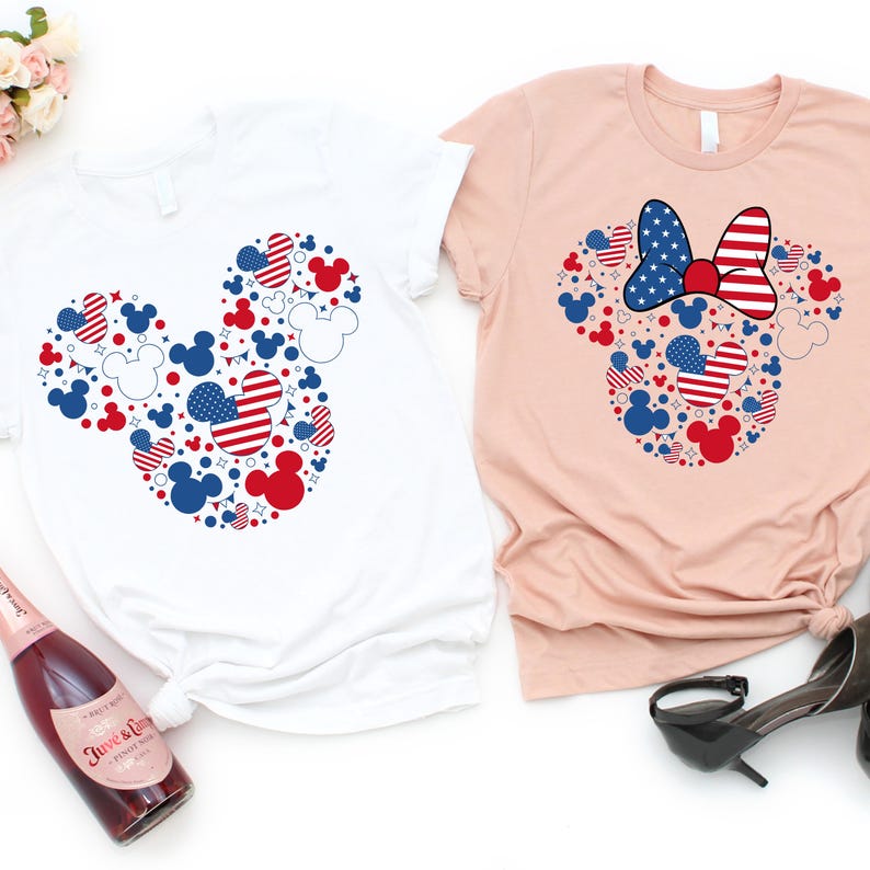 May include: Two t-shirts, one white and one light pink, each featuring a Mickey Mouse head design made of red, white, and blue shapes. The design incorporates stars, stripes, and Mickey Mouse silhouettes, celebrating a patriotic theme.