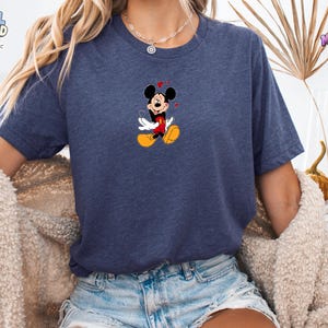 May include: A heather navy blue short-sleeve t-shirt featuring a centered graphic of Mickey Mouse with his eyes closed, wearing red shorts and yellow shoes, surrounded by small red hearts. The shirt has a crew neckline and a casual fit.