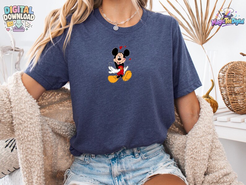May include: A heather navy blue short-sleeve t-shirt featuring a centered graphic of Mickey Mouse with his eyes closed, wearing red shorts and yellow shoes, surrounded by small red hearts. The shirt has a crew neckline and a casual fit.