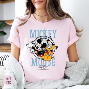 May include: A light pink t-shirt featuring a graphic of Mickey Mouse and Pluto inside a circular design with the words "Mickey Mouse" in light blue. The shirt has short sleeves.