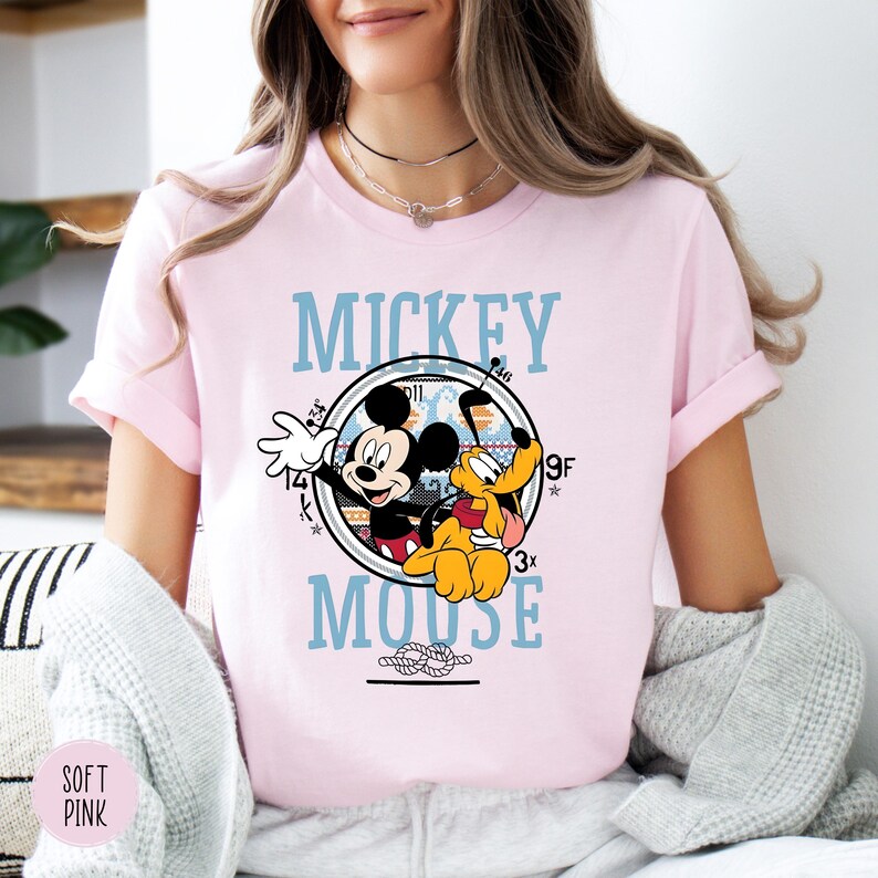 May include: A light pink t-shirt featuring a graphic of Mickey Mouse and Pluto inside a circular design with the words "Mickey Mouse" in light blue. The shirt has short sleeves.