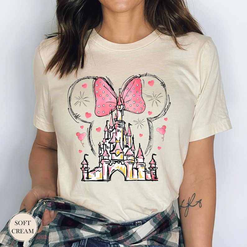 Puede incluir: Camiseta color crema con un dise&ntilde;o de castillo caprichoso, un lazo rosa y orejas de rat&oacute;n. El castillo tiene detalles rosas y amarillos, rodeado de corazones y estrellas. El texto "SOFT CREAM" est&aacute; en la parte inferior izquierda.