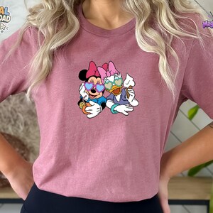 May include: A mauve short-sleeved t-shirt featuring a graphic print of Minnie Mouse and Daisy Duck wearing heart-shaped sunglasses. Minnie holds an orange drink with a straw, while Daisy holds a white ice cream cone. The design is centered on the chest.