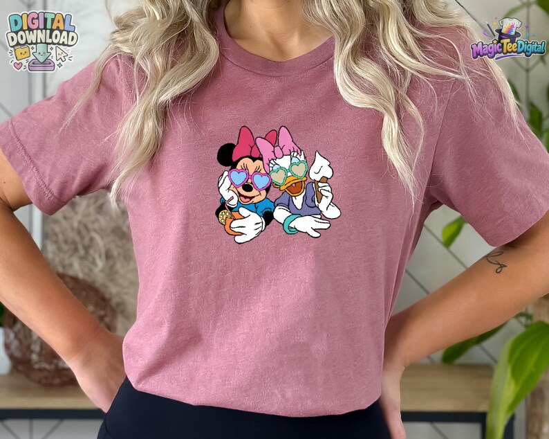 May include: A mauve short-sleeved t-shirt featuring a graphic print of Minnie Mouse and Daisy Duck wearing heart-shaped sunglasses. Minnie holds an orange drink with a straw, while Daisy holds a white ice cream cone. The design is centered on the chest.