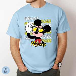 May include: Light blue t-shirt featuring a graphic of Mickey Mouse wearing yellow sunglasses and holding them up with his white gloved hand. The word "Mickey" is in yellow, and the word "MOUSE" is repeated in the background.