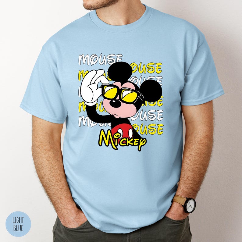 May include: Light blue t-shirt featuring a graphic of Mickey Mouse wearing yellow sunglasses and holding them up with his white gloved hand. The word "Mickey" is in yellow, and the word "MOUSE" is repeated in the background.