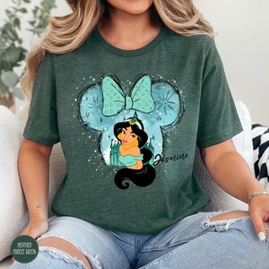 Puede incluir: Camiseta verde bosque jaspeado con un dise&ntilde;o de la Princesa Jasmine dentro de una silueta de Mickey Mouse y un lazo. El dise&ntilde;o incluye el nombre "Jasmine" en cursiva. La camiseta es de un material suave.