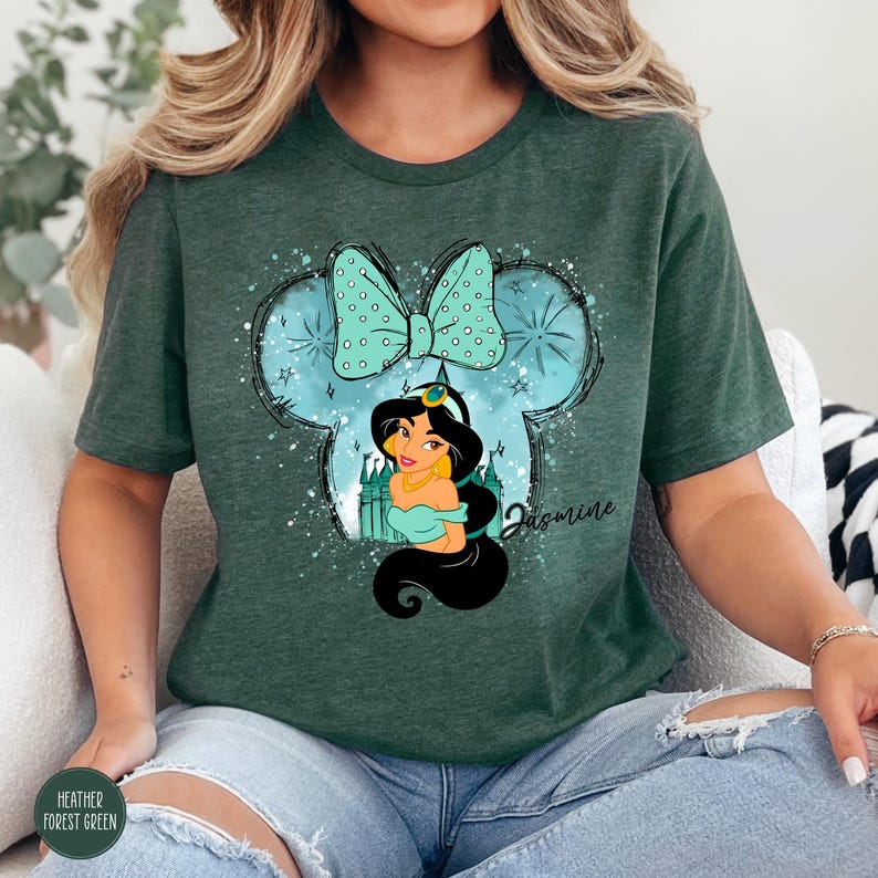 Puede incluir: Camiseta verde bosque jaspeado con un dise&ntilde;o de la Princesa Jasmine dentro de una silueta de Mickey Mouse y un lazo. El dise&ntilde;o incluye el nombre "Jasmine" en cursiva. La camiseta es de un material suave.