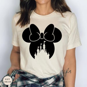May include: Cream-colored t-shirt featuring a black silhouette of a bow with a castle design inside. The text "SOFT CREAM" is printed on the bottom left of the shirt. The shirt is made of a soft material.